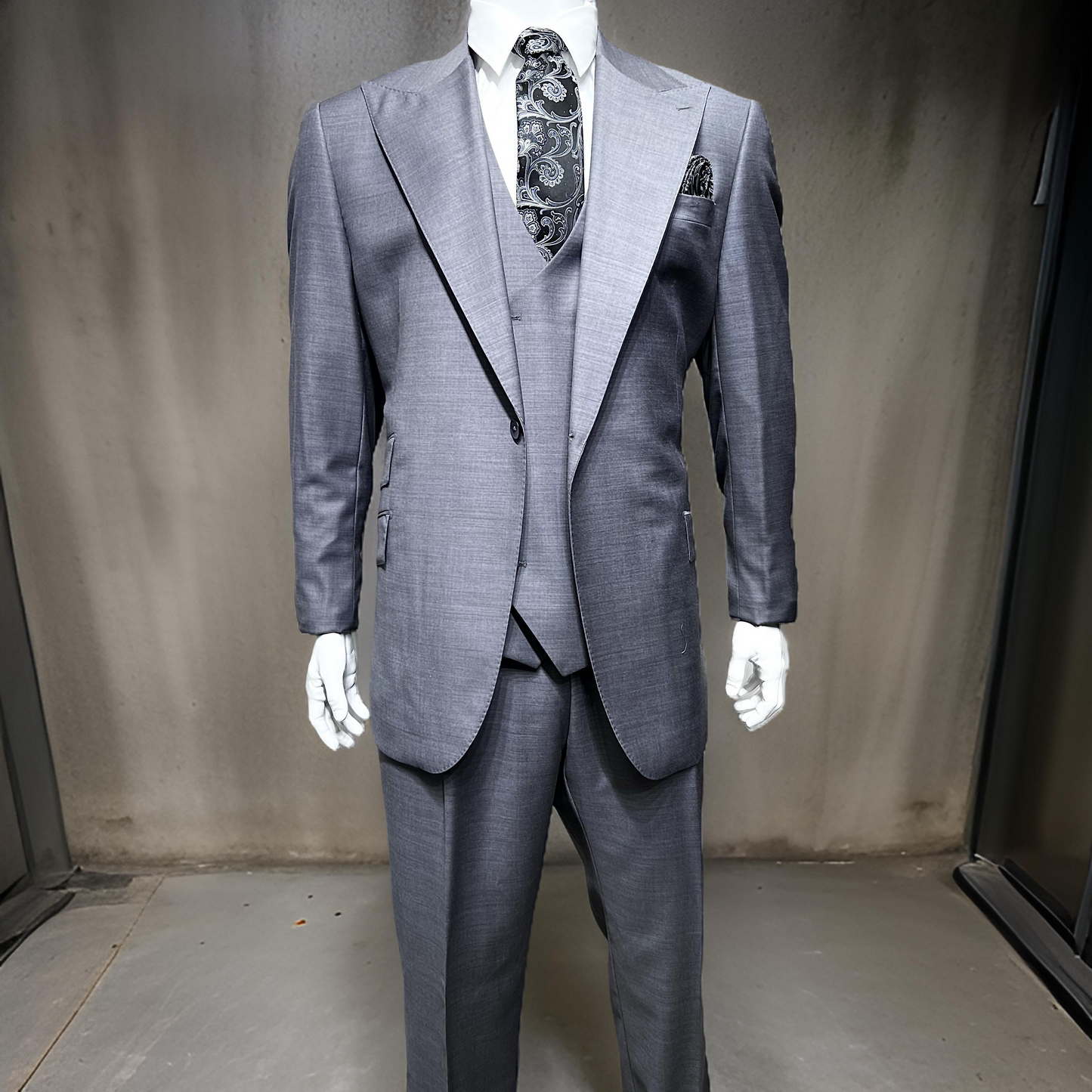 A mannequin elegantly dressed in the Tiglio Rosso Orvietto Solid Grey E09063/26 Single Pleat three-piece suit, crafted from 100% wool and complemented by a patterned tie and pocket square, stands against a neutral textured background that perfectly highlights the sophistication of Tiglio Rosso suits.