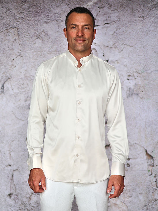 A man stands against a textured light gray wall, wearing the Manzini IN-101 Regular Collar Solid Color Satin Shirt in cream and white pants, facing forward with a neutral expression.