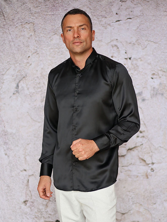 A man wears the MANZINI INR-102 Banded Collar Solid Color Satin Shirt in Black by Manzini with white pants, standing before a rough, light-colored textured wall.