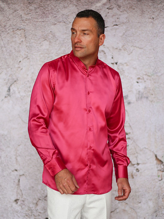 A man wears the Manzini INR-102 Banded Collar Solid Color Satin Shirt in fuchsia with white pants, standing before a textured light-colored wall.