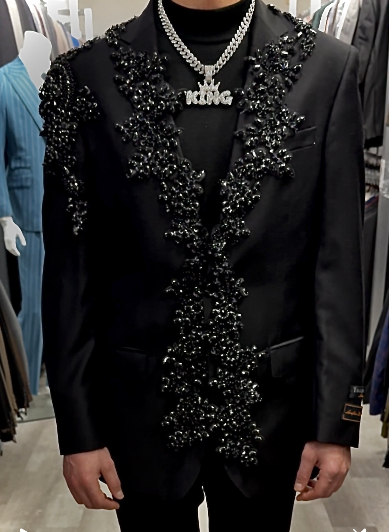 A person in a Unique Design Menswear Custom Prom Suit (VARIETY COLORS) with intricate beaded designs and a thick chain necklace stands in a clothing store, possibly ready to ask about the latest fashion trends.