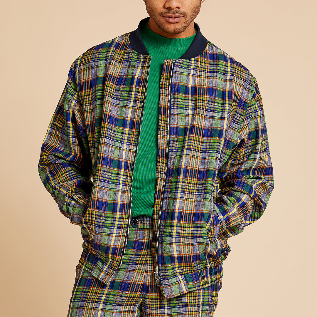 A person wearing a green shirt and the Inserch Linen Madras Bomber Jacket JS259-66 Multi, a colorful zip-up by Inserch with a dark ribbed collar, shown from the chest up against a beige background.