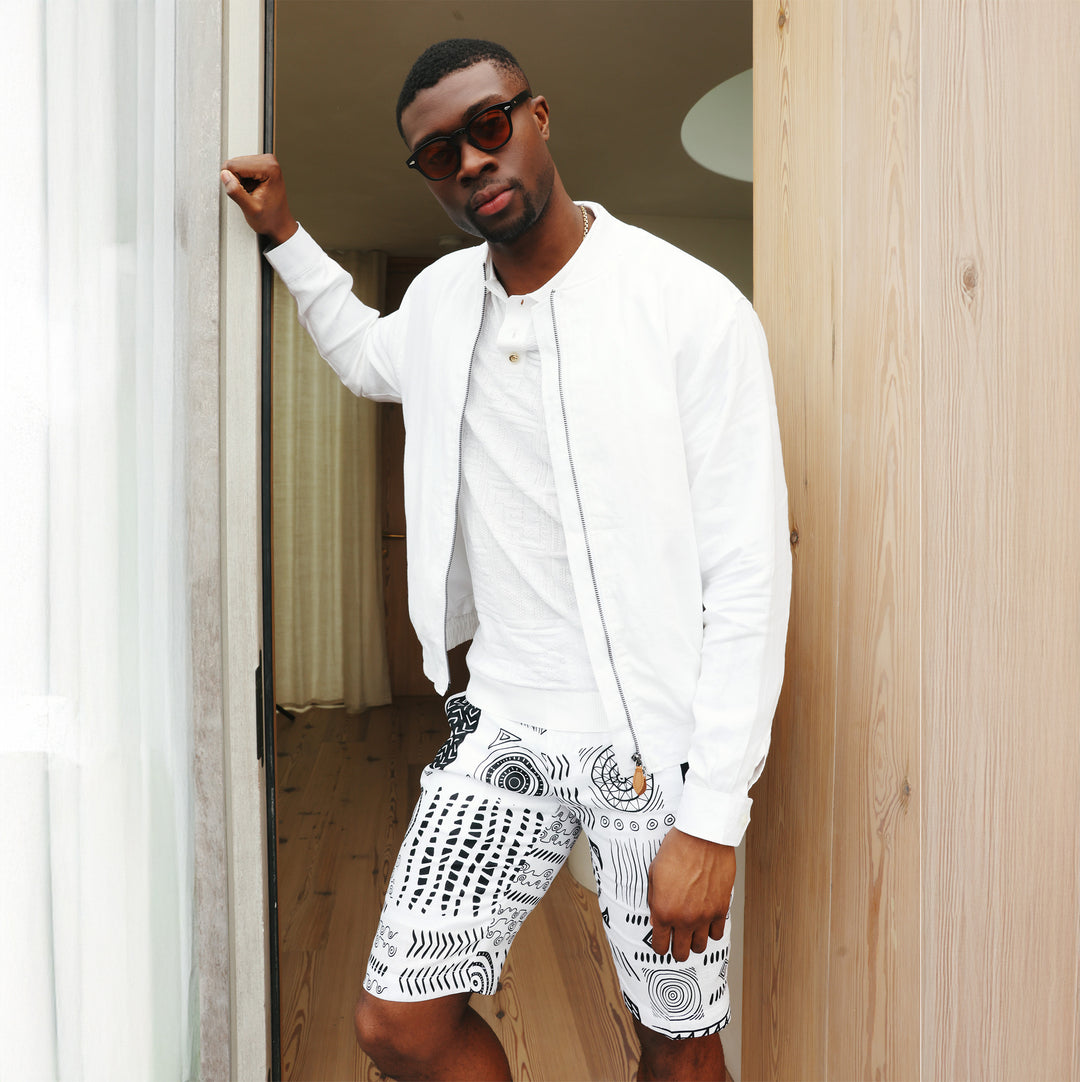 A man wearing the Inserch Premium Linen Short Zip Jacket JS660-02 White by Inserch, a white shirt, patterned white shorts, and sunglasses stands in a doorway, holding the door frame with one hand.