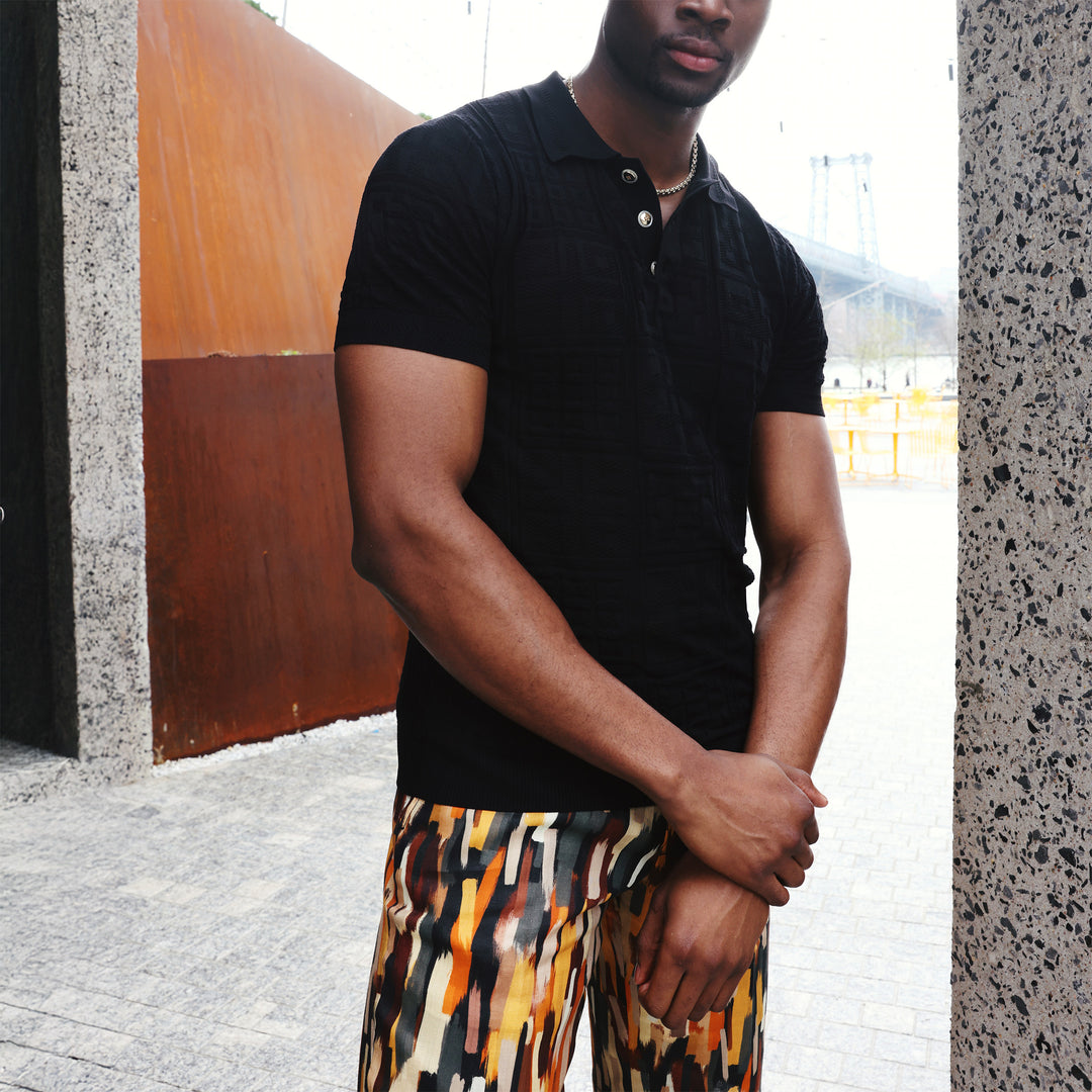 A man wears the Inserch SS Knit Polo KS001-00001 Black, a textured geometric knit short-sleeve shirt with novelty buttons by Inserch, paired with red pants against a beige background.
