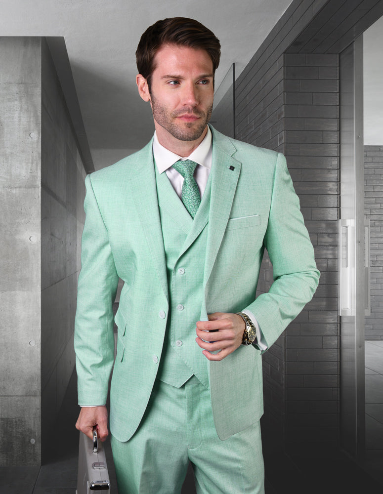 A man in the STATEMENT CLOTHING | LANZO-MINT slim fit suit by Statement Clothing, with a white shirt and green tie, stands indoors holding a silver briefcase against modern gray walls.