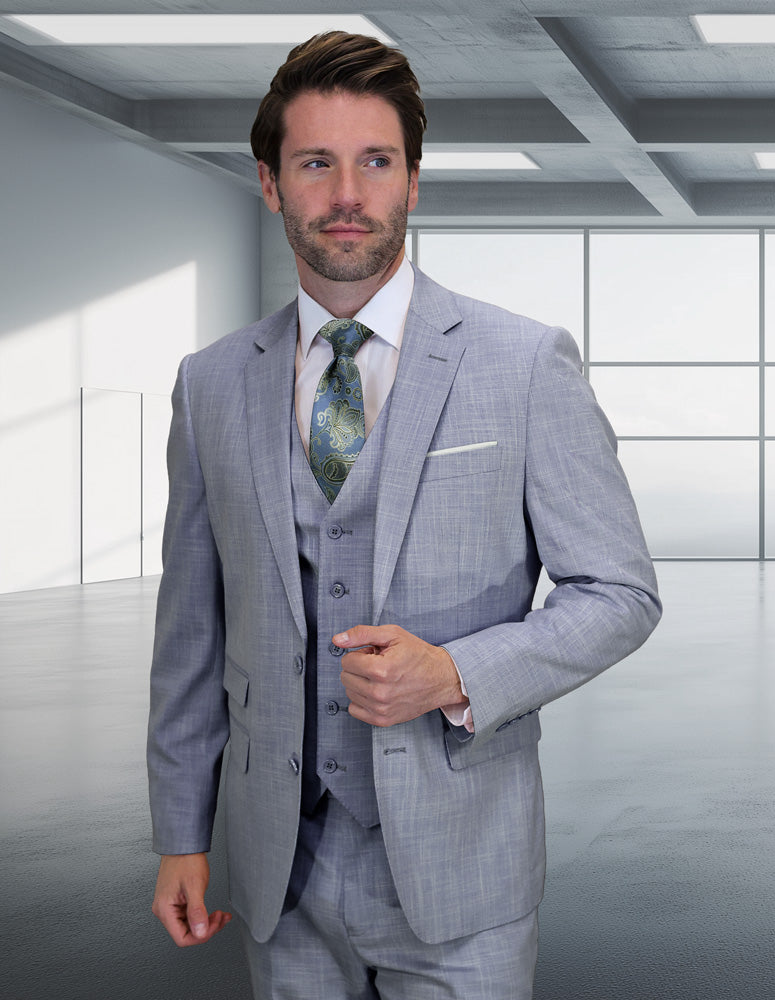 A man in the STATEMENT CLOTHING | LARZO-SKY BLUE suit by Statement Clothing, paired with a patterned tie, stands in a modern, empty room with large windows and a grid ceiling.