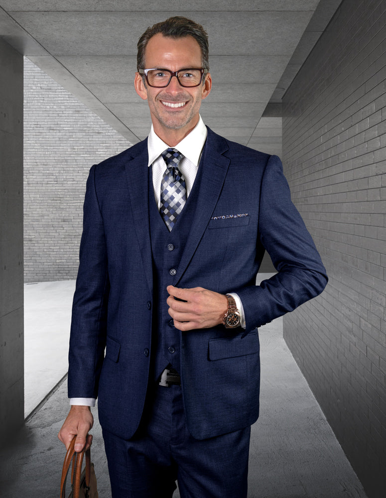 A man in a STATEMENT CLOTHING | LAZARO-NAVY suit by Statement Clothing, wearing glasses, stands in a modern concrete hallway, holding a brown briefcase and smiling at the camera.