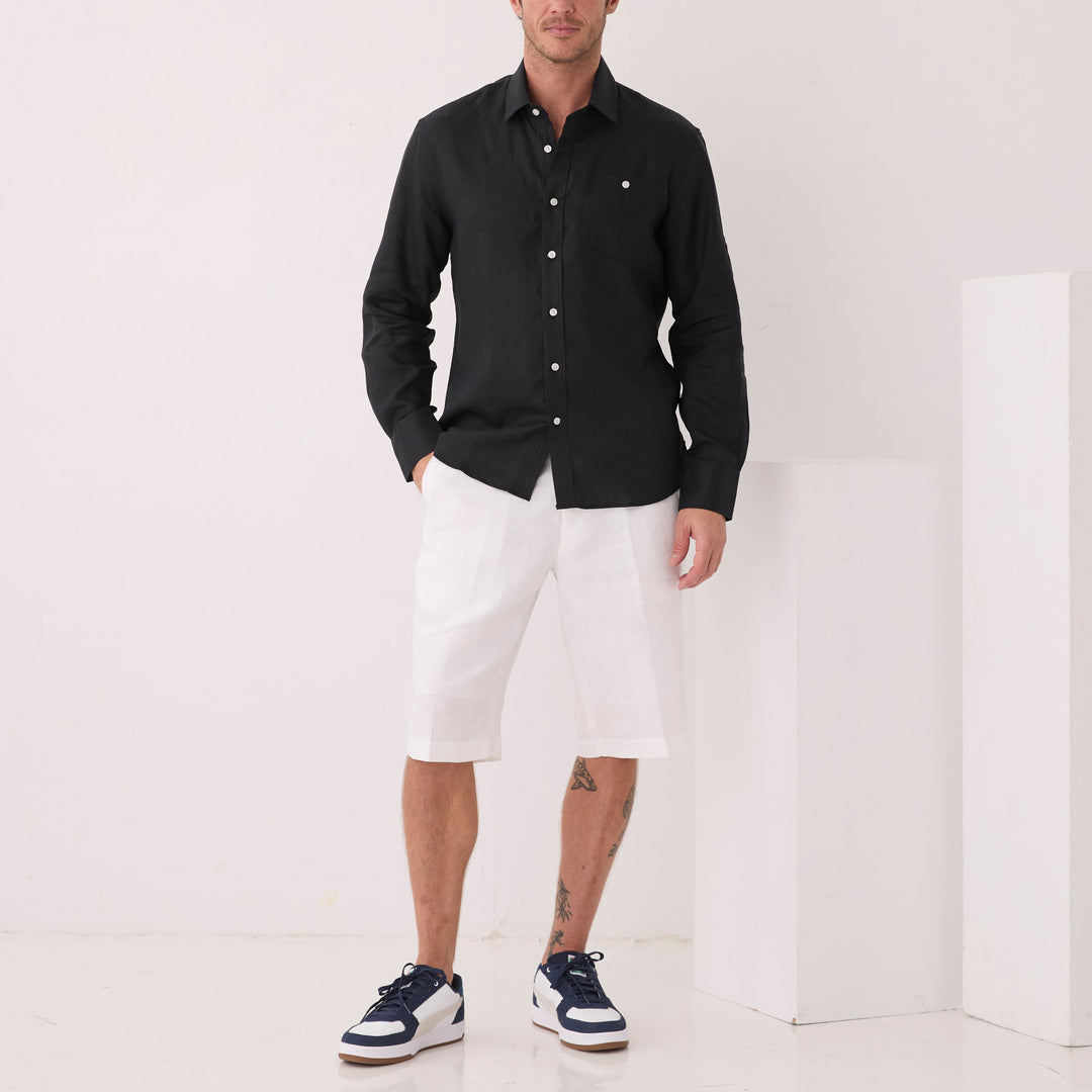 A man wears the Inserch Long Sleeve Modern Fit Linen Shirt in black, paired with white shorts and navy sneakers. He stands against a white background with geometric shapes, subtly showcasing the elegance of premium European flax used in the shirt's construction.