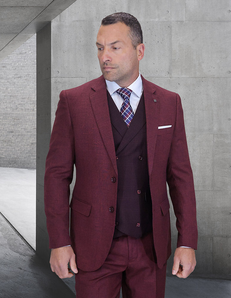 A man wears the STATEMENT CLOTHING | MARCO-1-BURGUNDY modern fit suit by Statement Clothing, paired with a patterned tie, standing indoors against a concrete wall and looking to his left with a neutral expression.