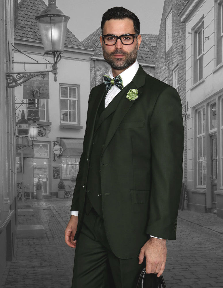 A man wearing the STATEMENT CLOTHING | MESSINA-OLIVE Italian wool suit by Statement Clothing, featuring pleated pants, a bow tie, and boutonnière, stands on a cobblestone street with historic buildings in the background.