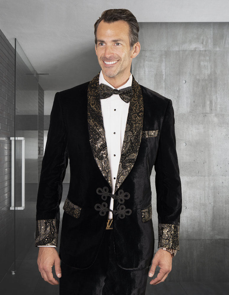 Man wearing the STATEMENT CLOTHING | MIRAGE-BLACK slim fit tuxedo by Statement Clothing, featuring ornate gold detailing, velvet vest, and bow tie, standing indoors against a modern minimalist background.