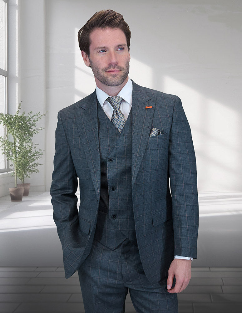 A man in the STATEMENT CLOTHING | MONTE-OLIVE gray plaid Italian wool suit by Statement Clothing, with a white shirt and indigo-patterned tie, stands indoors by a window and a potted plant.