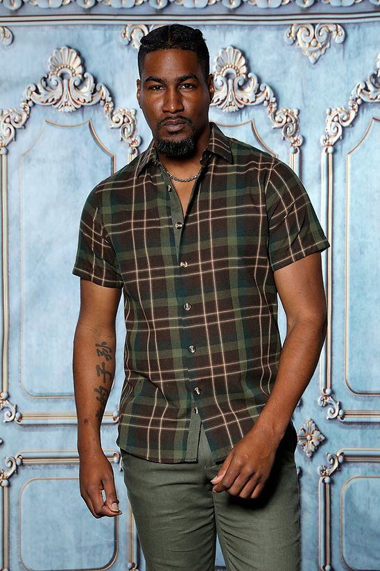 A man in a Manzini MANZINI MZL-302 2PC Plaid Linen Shirt Set in Kelly Green stands before an ornate blue decorative wall, paired with gray pants for a casual look.