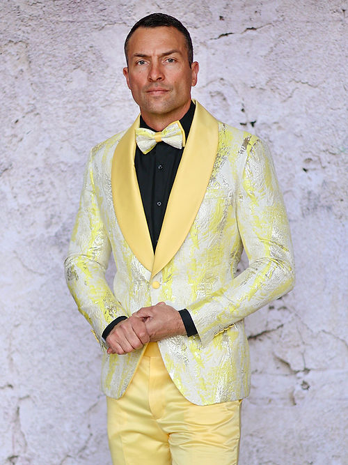Against a textured, light wall, a person wears the Insomnia MANZINI MZS-552 Textured Metallic Tuxedo Suit featuring a yellow jacket with shawl lapel and matching bow tie.