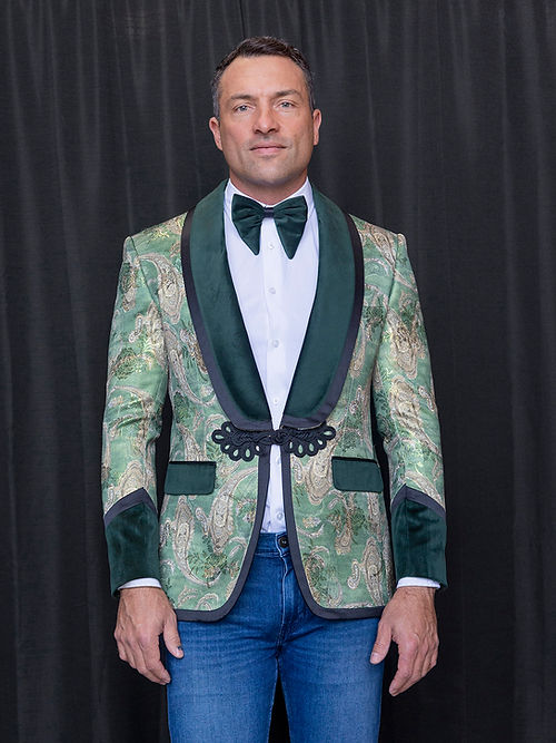 A man stands before a black curtain wearing a Manzini MANZINI MZS-566 Men's Tailored-Fit Fancy Velvet Paisley Tuxedo Dinner Jacket in green with black trim, a white shirt, green bow tie, and blue jeans.