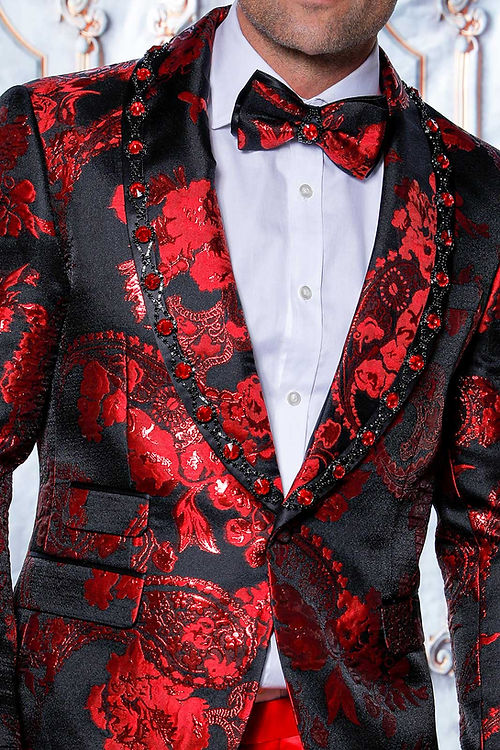 Man wearing a Manzini MZS-602 Rhinestone Shiny Metallic Floral Tuxedo Prom Red Jacket, styled with red pants and a bow tie, stands against an ornate backdrop.