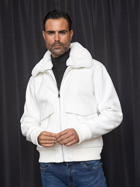 A man in an Insomnia MANZINI MZW-501 White Bomber Jacket Coat with a faux fur collar and blue jeans stands against a dark curtain backdrop.