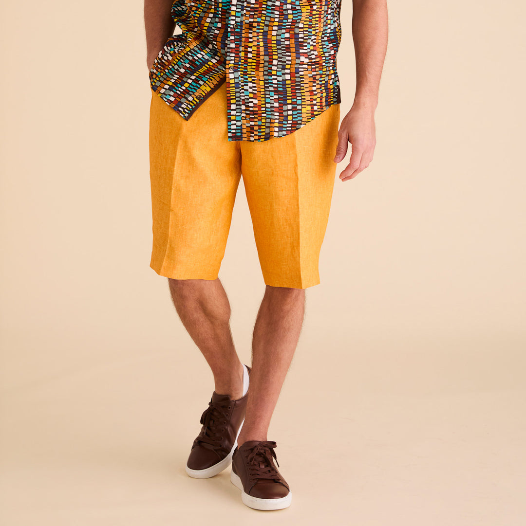 A person wearing a colorful patterned shirt, Inserch Premium Linen Flat Front Shorts in Sunburst, and brown sneakers stands against a neutral background.