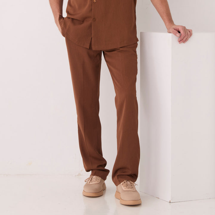 A person wears Inserch Crepe Flat Front Pants P9356-00064 Mocha, a matching brown shirt, and beige shoes, standing with one hand on a white block against a plain background.