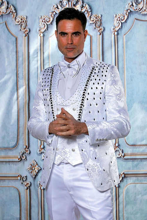 A man in an Insomnia MANZINI PLATINUM 3-Piece Slim Fit Rhinestone Satin Floral White/Black Tuxedo Suit stands with hands clasped against an ornate blue and silver decorative background.