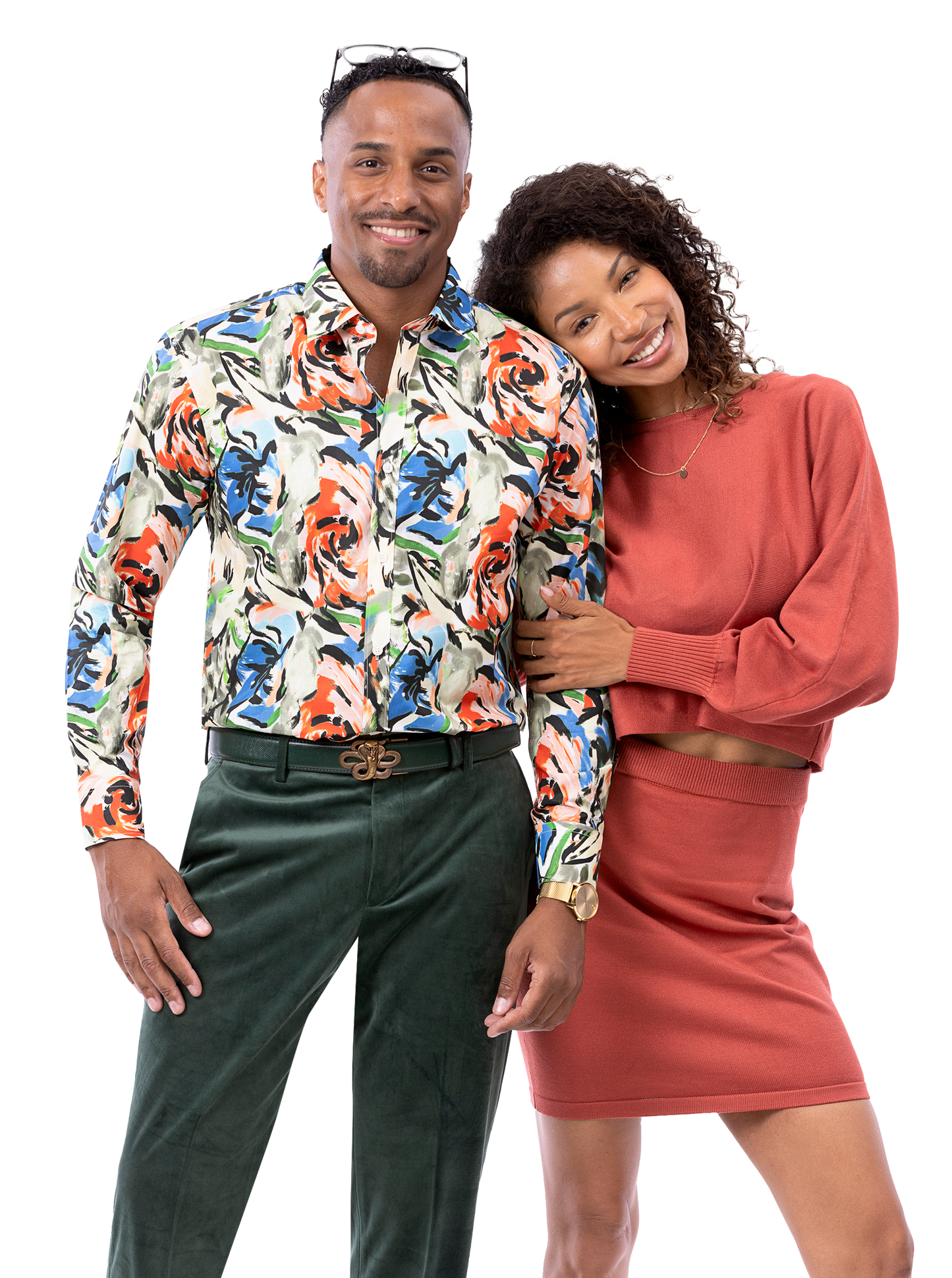 A man wearing an EJ SAMUEL floral shirt paired with EJ Samuel Hunter Pants PV01 smiles beside a woman in a rust-colored outfit, who leans on his shoulder as they both strike a friendly pose.