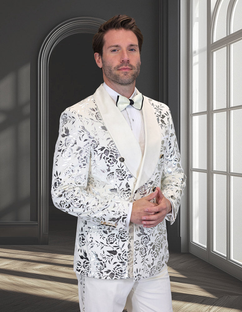 A man stands in a bright room wearing the STATEMENT CLOTHING | RENESANSE-IVORY tuxedo from Statement Clothing, featuring a white shawl lapel, floral lace pattern, crisp white shirt, and matching bow tie.