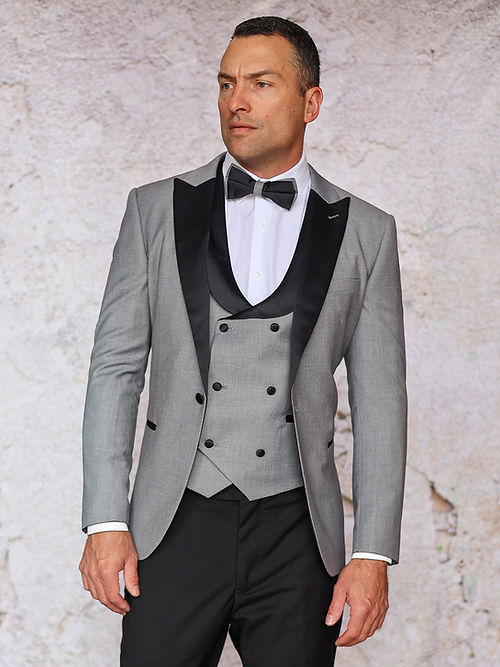 A man stands against a light textured wall wearing the Insomnia MANZINI ROME 3-Piece Slim Fit Stretch Spandex Grey Tuxedo Suit with black lapels, white shirt, black bow tie, grey waistcoat, and black trousers.