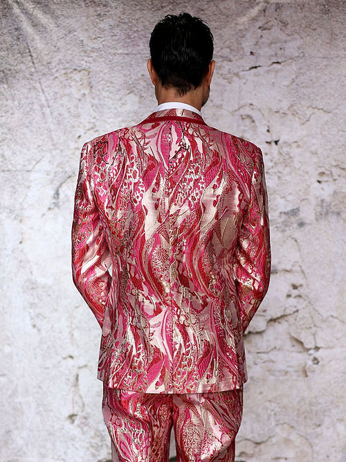 A man confidently stands against a textured light background in an Insomnia Manzini Santo 3-piece slim fit fuchsia tuxedo suit with a bright pink and gold metallic pattern, complemented by a matching bow tie.
