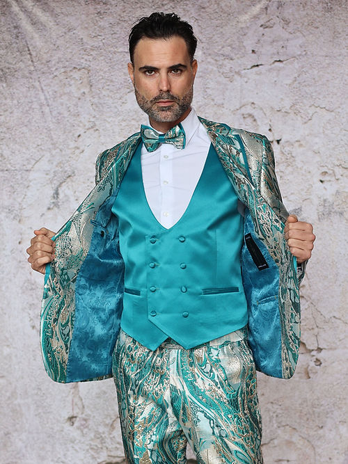 A man poses against a textured background in an Insomnia MANZINI SANTO turquoise tuxedo suit, featuring a fancy metallic pattern and bow tie, looking down.