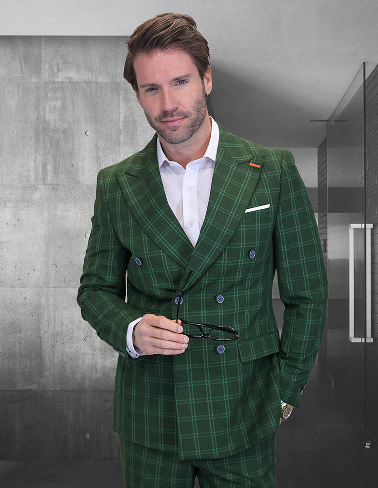 A man wears the STATEMENT CLOTHING | SD-103-OLIVE green plaid double-breasted Italian wool suit by Statement Clothing, holding eyeglasses in a modern minimalist interior with concrete walls and glass doors.