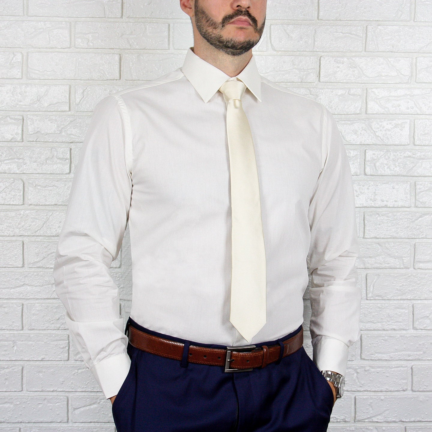 An individual in an ARTURO Slim Fit Long Sleeve Ivory Dress Shirt and a cream tie stands against a light brick wall, wearing navy pants with a hint of spandex and a brown belt.