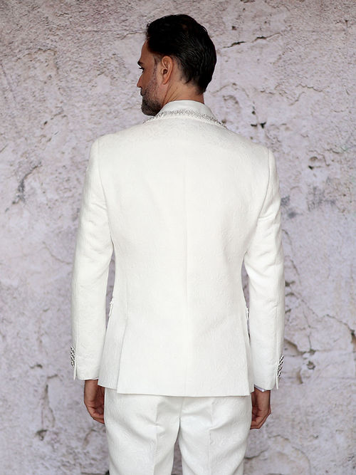 A man stands against a textured light background wearing the Insomnia MANZINI SOFIA 3-piece cream tuxedo suit, featuring a slim fit with satin shawl lapel and bow tie, and adorned with shiny rhinestone trim on the floral texture.