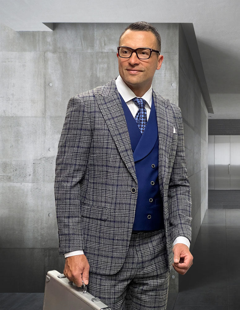A man in the STATEMENT CLOTHING | SORENTO-GRAY suit by Statement Clothing, with a blue vest, white shirt, and patterned tie, holds a silver briefcase in a sleek gray concrete hallway.