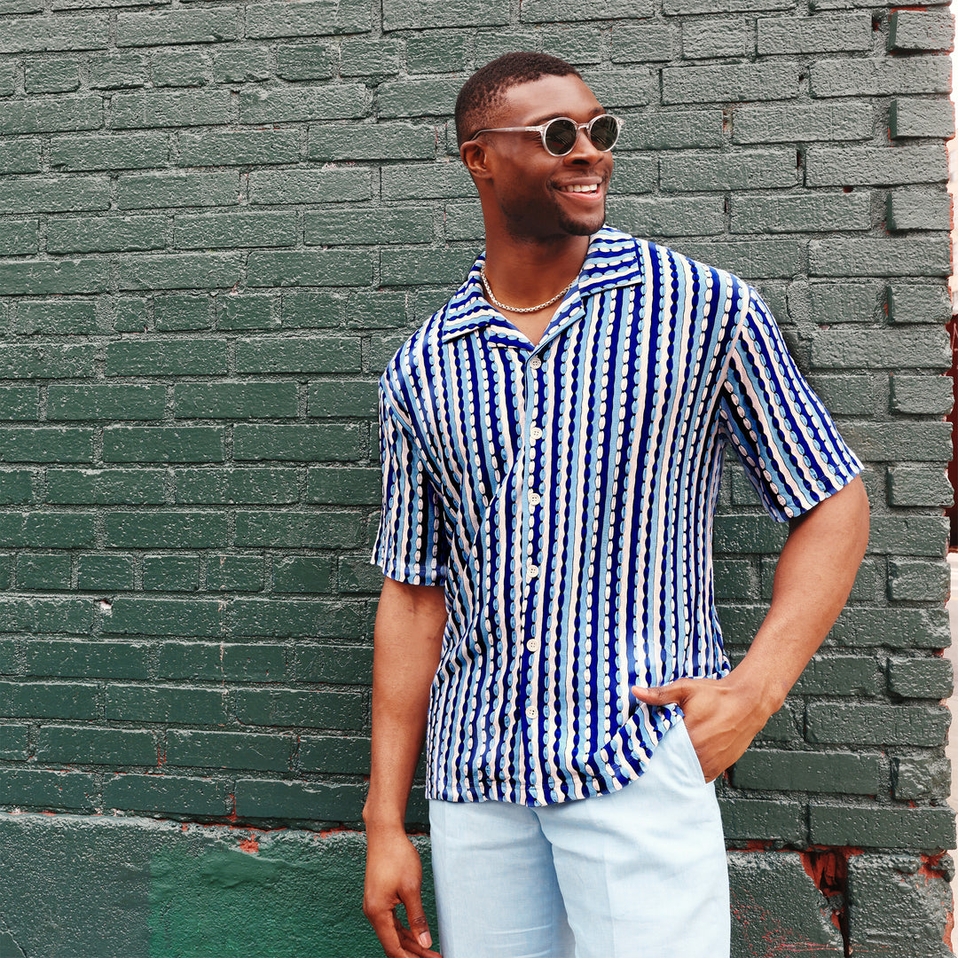 A man wearing the Inserch SS Knit Camp Collar Shirt SS020-00181 River Blue by Inserch, featuring blue, white, and black vertical stripes, with white pants, stands against a plain beige background.
