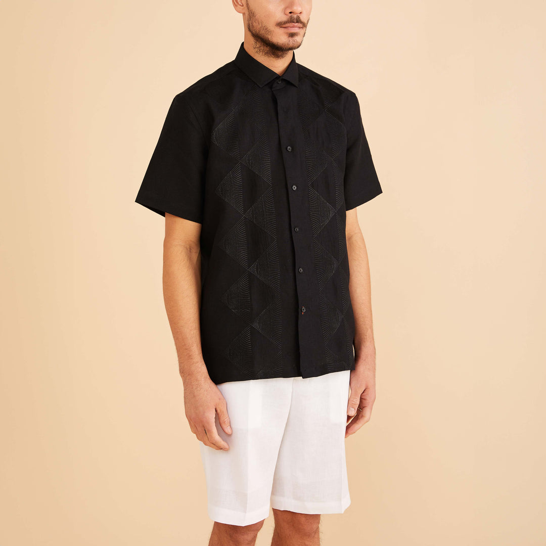 A man wearing the Inserch SS Linen Embroidered Shirt SS126-00001 in black pairs it with white shorts against a beige background.