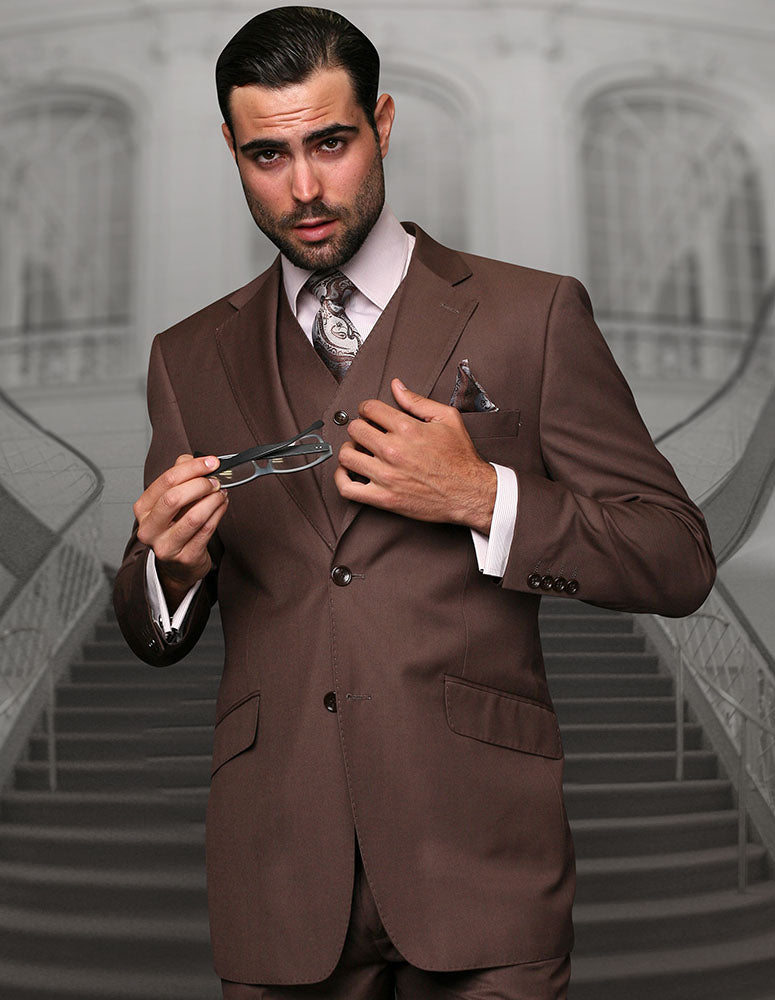 A man in a modern fit chestnut Italian wool STATEMENT CLOTHING | STZV-102-COCO suit by Statement Clothing holds eyeglasses while standing before a blurred staircase background.