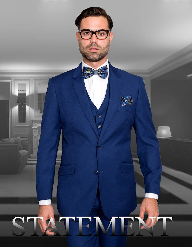 A man in a blue STATEMENT CLOTHING | STZV-102-SAPPHIRE suit by Statement Clothing, with a bow tie and glasses, stands confidently in a modern room. The word "STATEMENT" is displayed at the bottom.