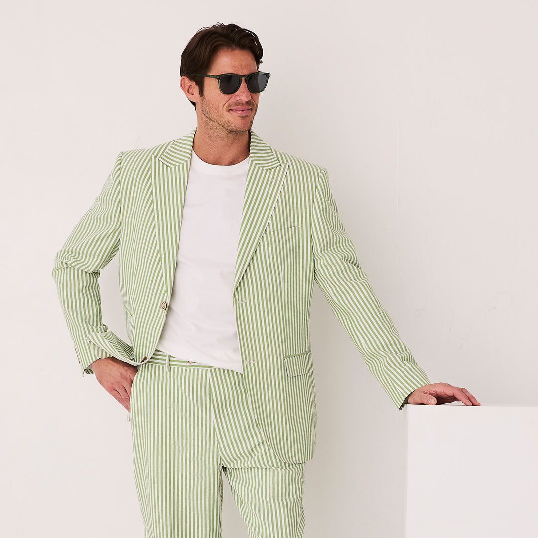 A person confidently stands, wearing the Inserch Seersucker Stripe Blazer SU660155-00018 in Kiwi over a crisp white shirt and sunglasses, with one hand resting on a white surface.