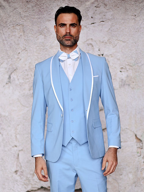 A man wearing the Insomnia MANZINI SUNSET Slim Fit 3-Piece Sky White Tuxedo Suit with a reversible vest and white bow tie stands against a textured beige wall.