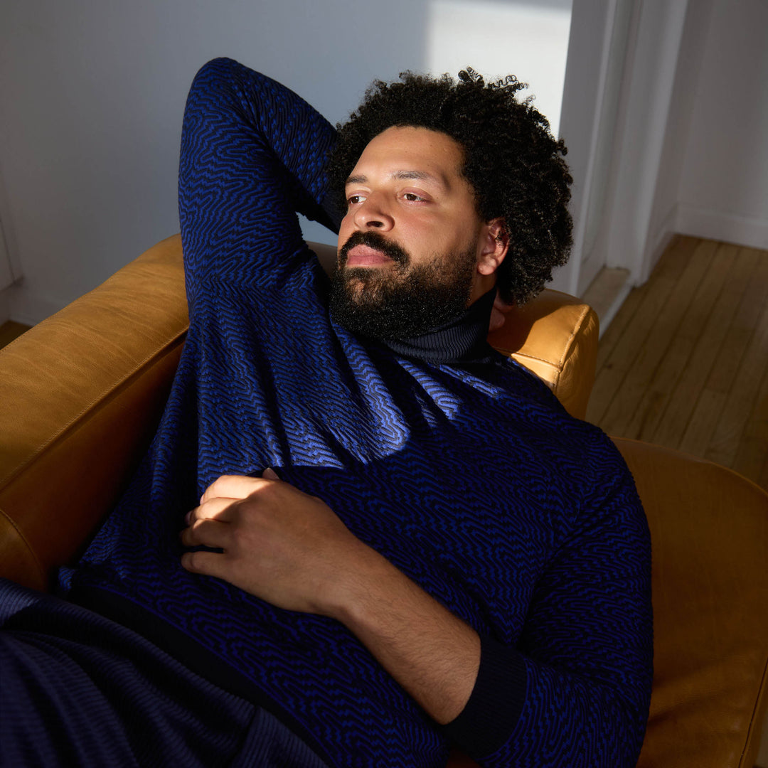 A man wearing the Inserch Intarsia Turtleneck Sweater SW304-00036 Ink by Inserch and dark blue pants stands against a plain beige background.