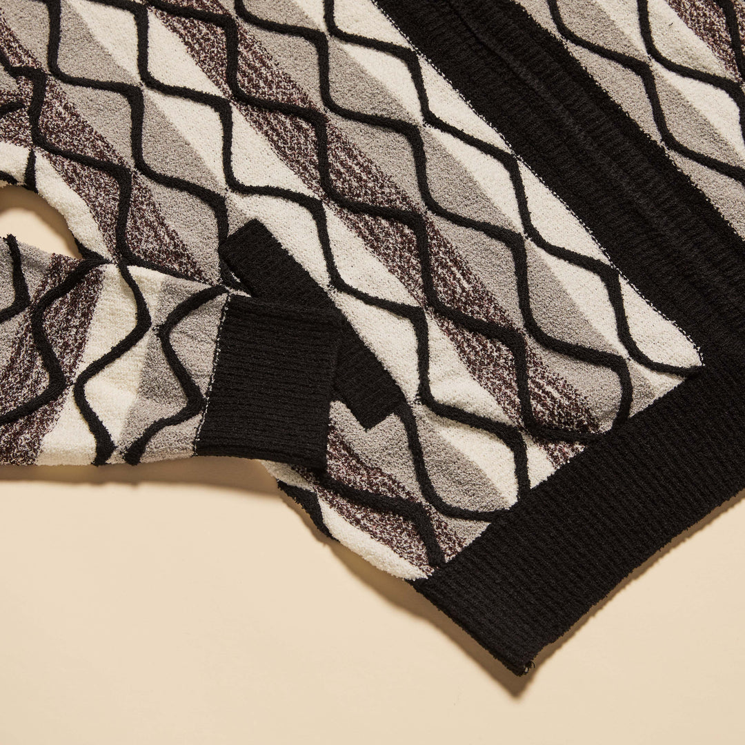 A man stands against a beige background wearing the Inserch Bouclé Yarn Graphic Full Zip Sweater (SW614-00001) in black, paired with a light gray turtleneck, black pants, and dark shoes.