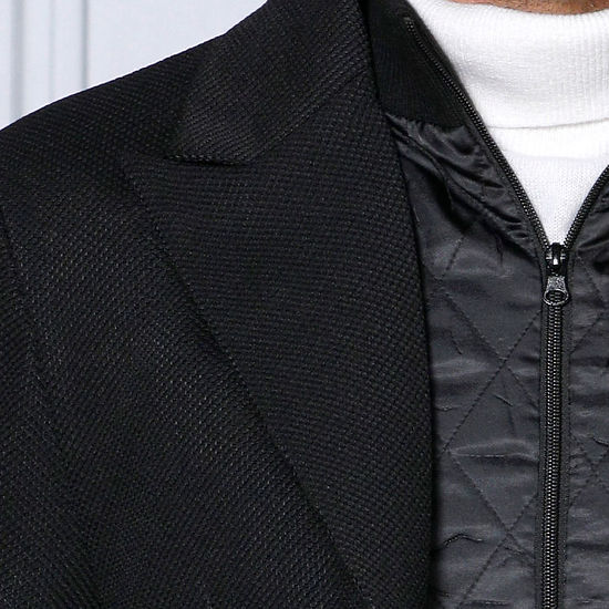 A man with short hair wears a MANZINI SWITZERLAND Black Blazer Coat with a zip-out liner from Insomnia, tailored fit vest, and jeans. He stands before an ornately decorated light gray wall, holding the coat's lapels.