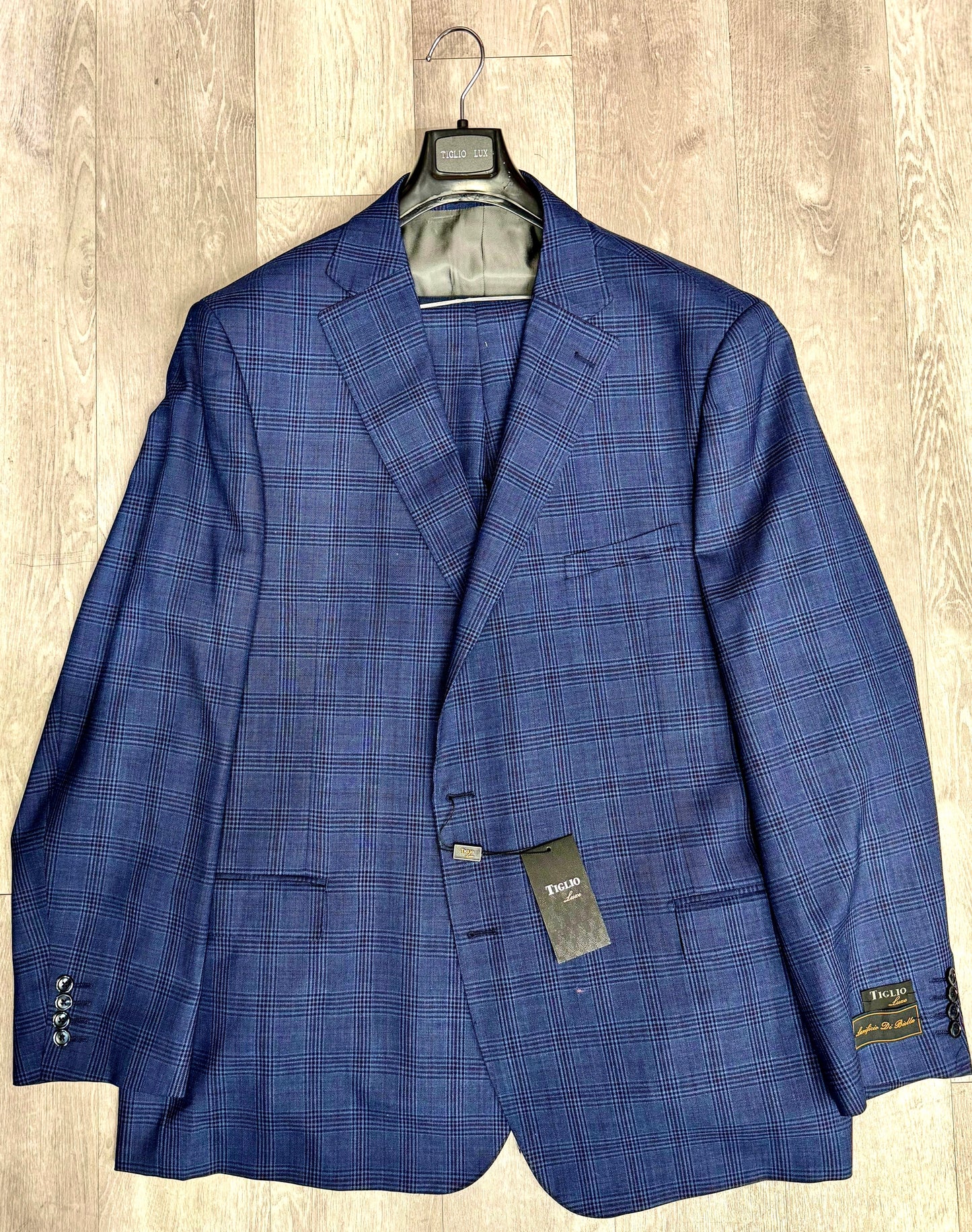 A Tiglio Luxe Dolcetto Modern Fit, Pure Wool Suit 332213/901 jacket in a blue checkered pattern, made from Italian fabrics, hangs elegantly on a wooden hanger with its tag attached, displayed on a wooden floor. This piece embodies timeless sartorial traditions.
