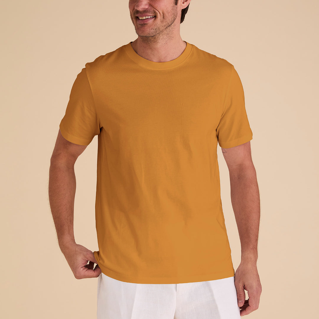 A man wearing the Inserch Short Sleeve Pima Cotton T-Shirt T301-00191 Sunburst and white pants stands against a beige background, smiling while holding his shirt's edge.