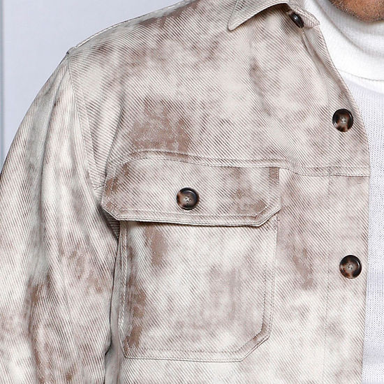 A man in a MANZINI VALENTINO Tan Soft Vegan Suede Button Sweater Shirt by Insomnia, paired with blue jeans, stands against a textured white wall.
