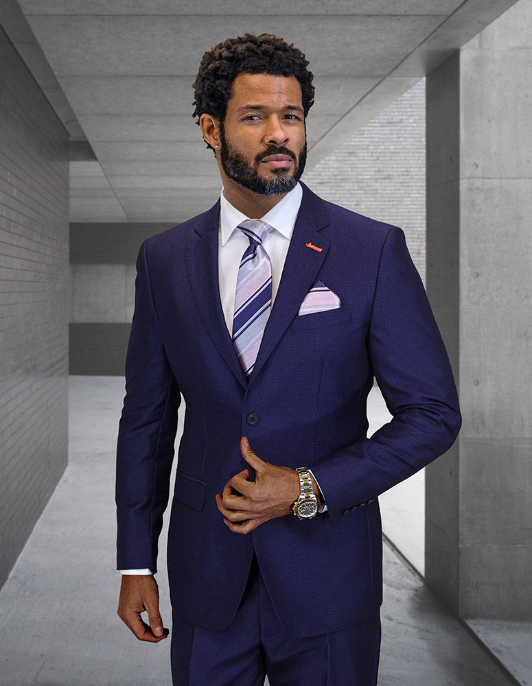 A man wearing the STATEMENT CLOTHING | VERNAZA-NAVY modern fit suit by Statement Clothing, paired with a white shirt and striped tie, stands in a sleek gray concrete hallway, looking ahead with one hand on his jacket.