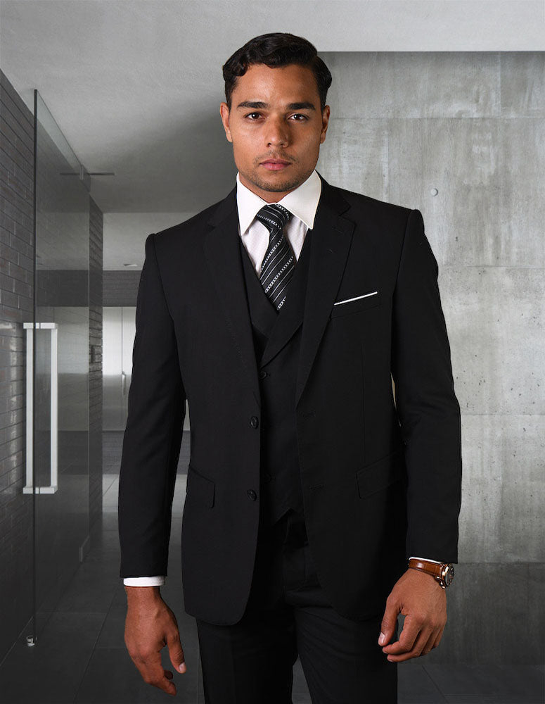 A man wearing the STATEMENT CLOTHING | ZARGALA-BLACK suit by Statement Clothing stands in a modern, minimalist office hallway with gray walls and glass doors.
