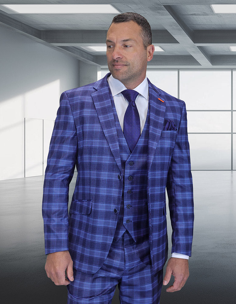 A man in the STATEMENT CLOTHING | ZILO-1-INDIGO modern fit suit by Statement Clothing, paired with a white shirt and blue tie, stands in a sleek room with large windows and a concrete ceiling.