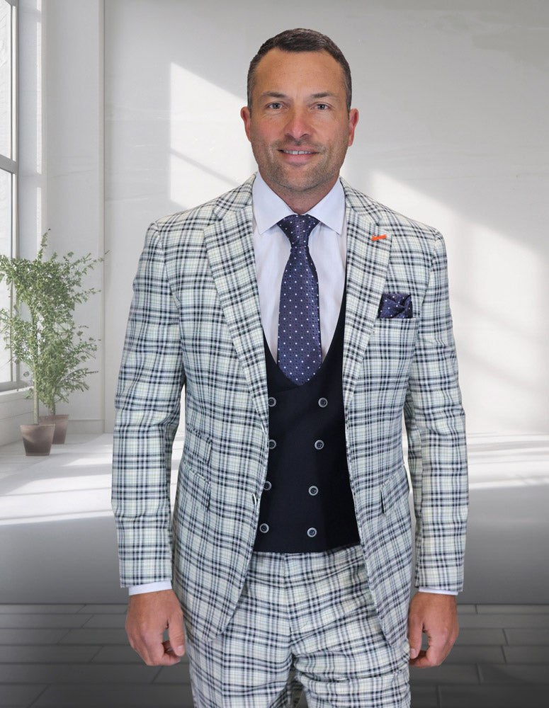 A man in a modern fit Statement Clothing ZILO-3-NATURAL Italian wool suit in white and blue plaid, with flat front pants, white shirt, and navy polka dot tie stands indoors by a potted plant as sunlight streams through large windows.