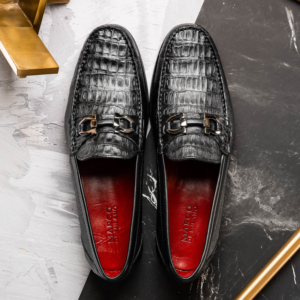 The Adro Black by Marco Di Milano are black textured leather loafers made from genuine exotics, with metal bit detailing and a red inner lining. One shoe stands upright while the other lies on its side to show the sole.